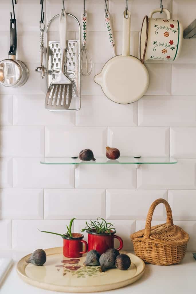 Charming kitchen scene with hanging utensils, red mugs, figs, and a wicker basket on white tile wall.